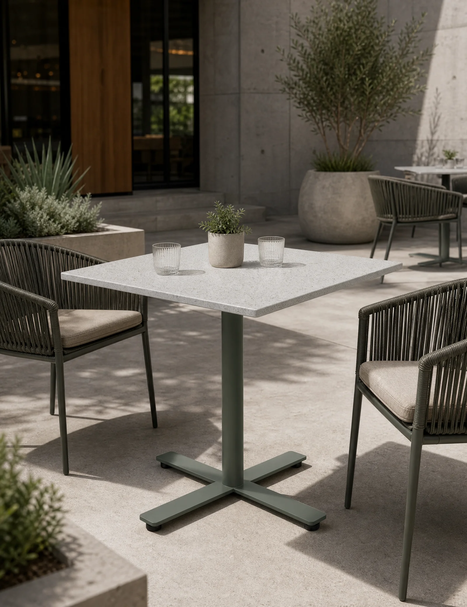 Element outdoor table with a stone-look top and X-base on a sunlit concrete patio, flanked by rope-woven cafe chairs